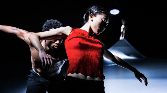 Twee dansers bewegen dicht langs elkaar op een donker podium. De danseres op de voorgrond draagt een felrode top en strekt haar armen uit, terwijl de danser achter haar een ondersteunende beweging maakt.