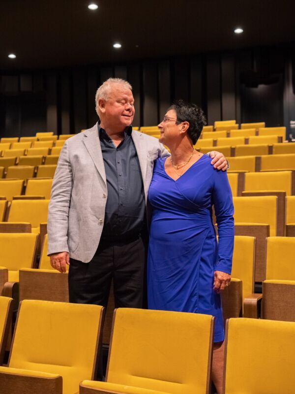 Hans Schriel and Ineke Roodenburg stand smiling arm in arm in a theatre auditorium with yellow seats.