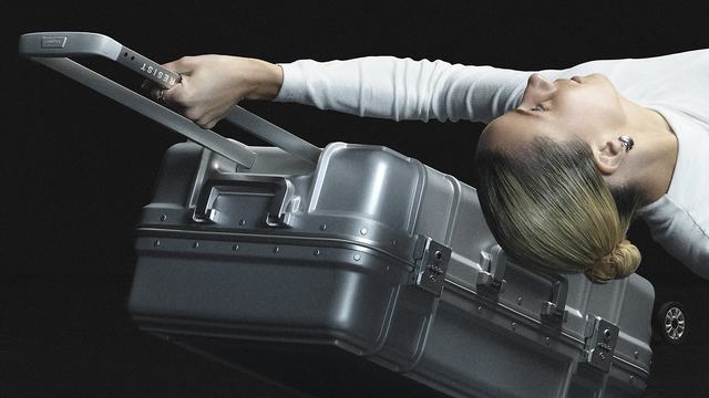 A dancer lies sideways on a dark stage, holding the extended handle of a silver suitcase. Her body forms a fluid, almost weightless line alongside the suitcase.