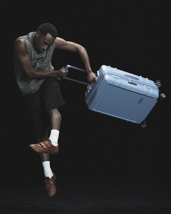 A dancer is captured mid-air against a dark background, holding a light blue suitcase by its extended handle.